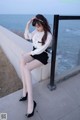 A woman sitting on a ledge by the ocean wearing a white blouse and black skirt.